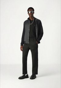 Melnā jaka ar sudraba pogām, melna T-krekls, zaļas bikses un melnas kurpes. Modelis nēsā sudraba saulesbrilles un ķēdes kaklarotu.