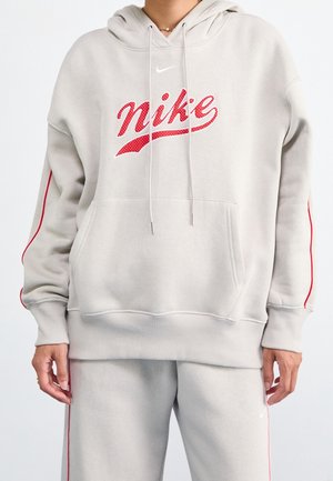 Person wearing light gray Nike hoodie and matching sweatpants with red stripes and red Nike logo on the chest and pant leg.