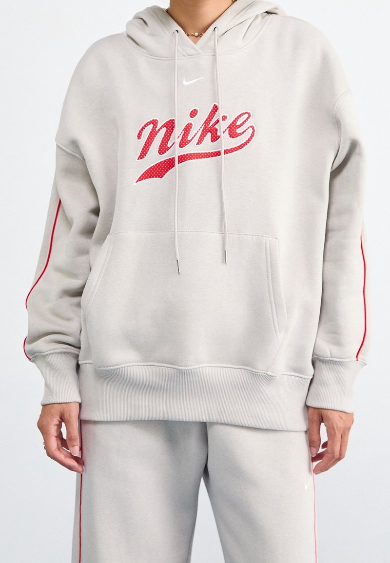 Personne portant un sweat à capuche Nike gris clair et un pantalon de survêtement assorti avec des bandes rouges et le logo Nike rouge sur la poitrine et la jambe du pantalon.