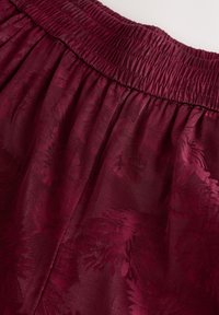 Burgundy-tyyppinen hame, jossa on smock-tyylinen vyötärö, ja joka esittelee teksturoitua lehtikuviota kiiltävässä pinnassa, korostaen sileää pinta-alaa ja laskosta.