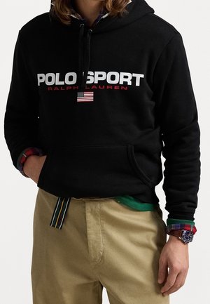 Personne portant un sweat à capuche Polo Sport Ralph Lauren noir avec un drapeau américain sous le logo, un pantalon beige et une montre au poignet gauche.