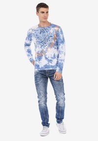 Langärmliges blaues Batikhemd mit orangefarbigem Grafixtyp; getragen mit hellblauen Jeans und weißen Sneakern. Es hat einen Rundhalsausschnitt.