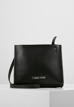Zwarte rechthoekige crossbodytas met dunne band en zilveren Calvin Klein-logo aan de voorkant, weergegeven op een witte ondergrond en een lichtgrijze achtergrond.