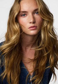 Lähivõte modellist laineliste, pikkade blondide juuste ja siniste silmadega. Tal on neutraalne ilme ja ta kannab lukuga teksajopet.