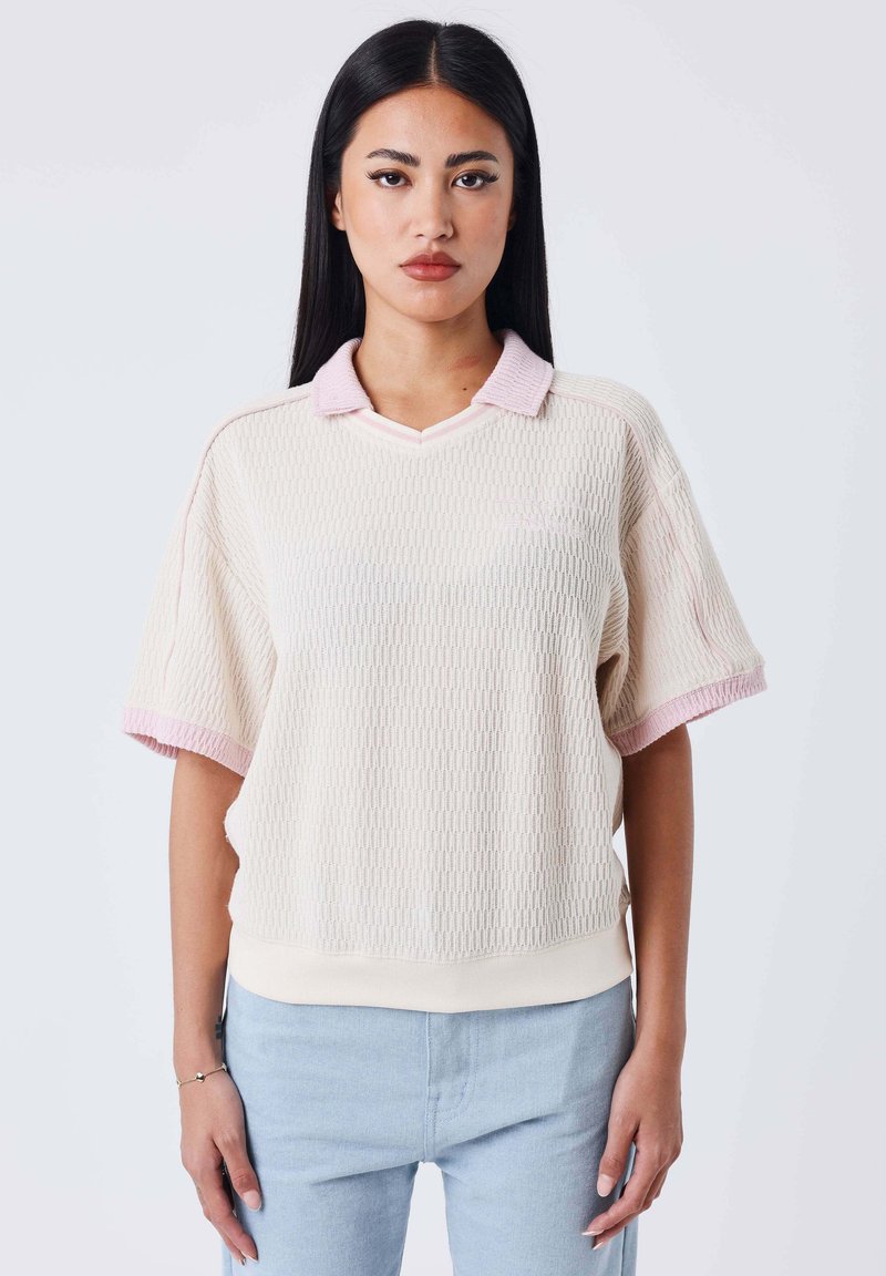 Jeune femme aux longs cheveux noirs portant un pull texturé rose pâle à manches courtes et un jean bleu clair, debout devant un fond uni.