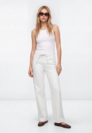 Woman wearing sleeveless white top, high-waist white pants with belt, brown flats, and sunglasses, standing against plain white background.