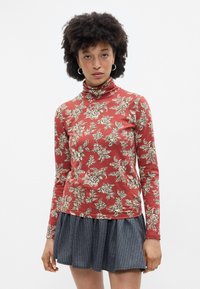 Top de gola alta de mangas compridas em vermelho com padrões de flores em creme, combinado com uma saia plissada cinza riscada. Textura suave por todo o conjunto.