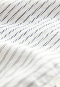 Baumwollstoff mit weißem Hintergrund zeigt dünne, gewellte blaue Streifen. Die Textur wirkt weich und glatt, mit sichtbaren Falten und Drapierungen.
