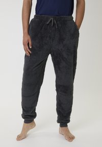 Personne portant un pantalon en polaire noir et moelleux avec des poignets élastiques et une taille à cordon, debout pieds nus contre un fond uni.