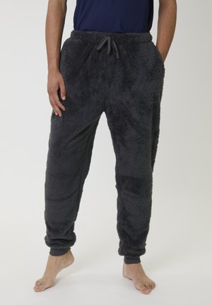 Persoon in zwarte donzige fleece broeken met elastische boorden en een koord in de taille, staat blootsvoets tegen een effen achtergrond.