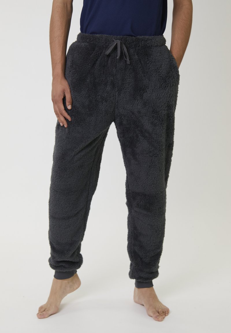 Personne portant un pantalon en polaire noir et moelleux avec des poignets élastiques et une taille à cordon, debout pieds nus contre un fond uni.