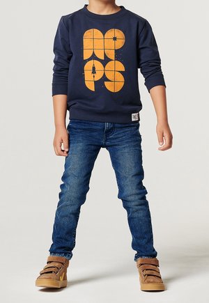 Barn står iført en mørkeblå sweatshirt med orange rumtema grafik, blå jeans og brune velcrosneakers.