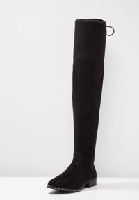 Bota de ante negra por encima de la rodilla con tacón bajo y tachuelas plateadas a lo largo del borde de la suela, sobre un fondo blanco liso.