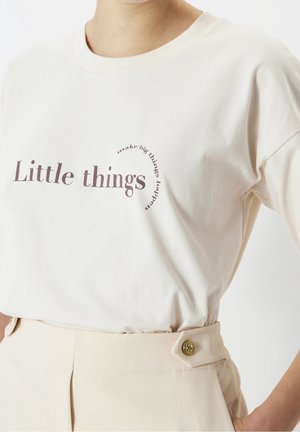Hvid T-shirt med lilla tekst "Little things" og "make big things happen" i en buet linje, kombineret med beige, højttaljede bukser med en knap.