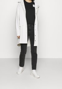 Persona vistiendo un abrigo de lluvia gris claro, camiseta negra, jeans gris oscuro y zapatillas blancas de pie contra un fondo liso.