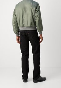 Veste bomber vert olive avec poignets et ceinture gris, associée à un jean noir. Présente une texture lisse et un design élégant. Chaussures noires visibles.