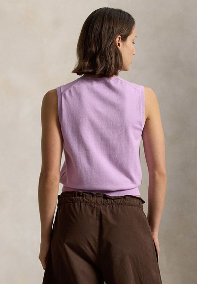 Γυναίκα με κοντά καστανά μαλλιά που φοράει μωβ αμάνικη μπλούζα και φαρδιά καφέ παντελόνια, όρθια με την πλάτη προς την κάμερα.