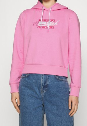 Osoba w różowej bluzie z kapturem z logo „Tommy New York Jeans” oraz niebieskich wysokich dżinsach, stojąca na jednolitym tle.