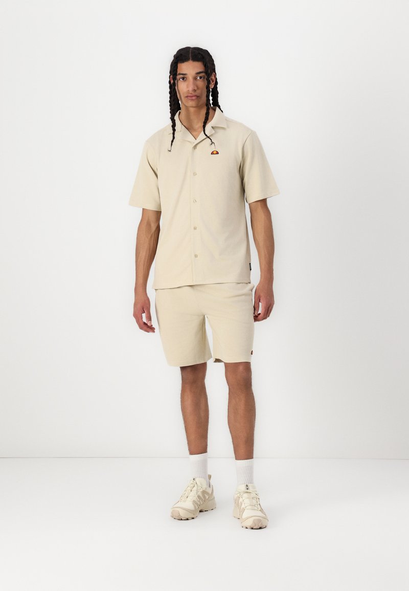 Camicia a maniche corte beige con bottoni e un piccolo logo colorato, abbinata a pantaloni corti beige e scarpe da ginnastica chiare.
