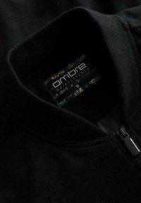 Czarna kurtka zapinana na zamek z wysokim kołnierzem, wykonana z teksturowanej tkaniny. Posiada metkę firmową wewnątrz, z napisem "ombre QUINTESSENCE," rozmiar M.