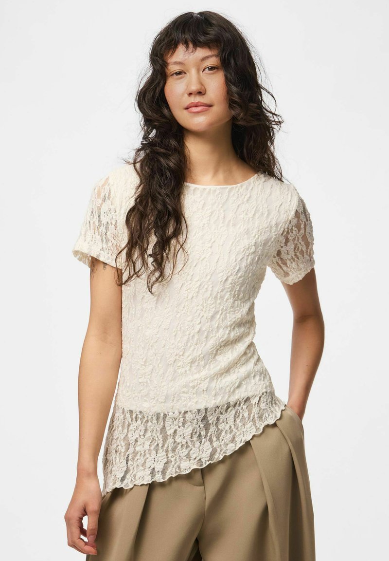 Jeune femme aux longs cheveux wavy foncés portant un haut en dentelle ivoire à manches courtes et un pantalon beige à jambes larges, debout devant un fond uni.