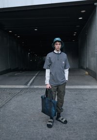 Gestreiftes T-Shirt, darunter ein weißes langärmeliges Shirt, olivgrüne Cargo-Hosen, dunkle Sandalen, türkisfarbene Bucket-Hat, hält eine dunkle Tragetasche, städtische Umgebung.