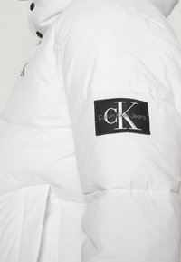 Giacca a vento bianca con texture liscia, caratterizzata da una toppa nera sulla manica con la scritta "Calvin Klein Jeans" e un logo. Bottoni a pressione visibili.