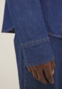 Abbigliamento in denim di un blu intenso, con una finitura leggermente strutturata, cuciture arancioni ben visibili e un design delle maniche con vestibilità rilassata.