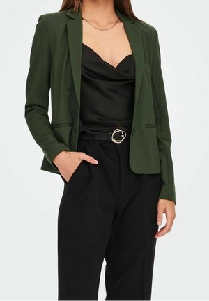 Femme portant un blazer vert foncé sur un caraco en satin noir, pantalon noir taille haute avec une ceinture noire avec boucle dorée.