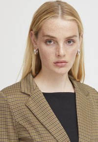 Brun och beige rutigt kavaj med en strukturerad slag, bärs över en svart topp. Slät tyg med skräddarsydd passform och minimal detaljering.