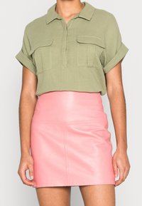 Blouse vert olive à manches courtes avec col à boutons et deux poches avant, associée à une mini-jupe en cuir rose taille haute.