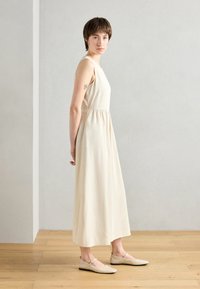 Light beige sleeveless dress with a fitted bodice and gathered waist, paired with nude flat shoes. Smooth texture, floor-length design.