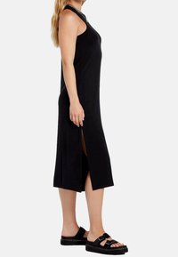 Robe noire sans manches avec fentes latérales, confectionnée en tissu lisse. Associée à des sandales noires à double boucle.