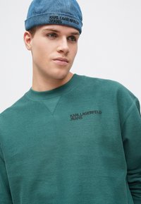 Tyrkysová mikina se žebrovanou strukturou a kulatým výstřihem, na hrudi s vyšitým nápisem "KARL LAGERFELD JEANS" v černé barvě. K tomu denimová čepice jako doplněk.