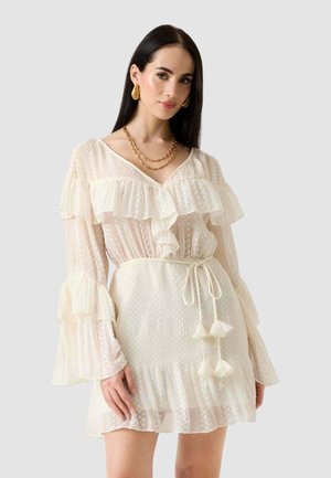 Frau mit langen dunklen Haaren, die ein cremefarbenes Kleid mit Rüschen und transparenten Ärmeln trägt, einen Taillengürtel, goldene Ohrringe und geschichtete goldene Halsketten.