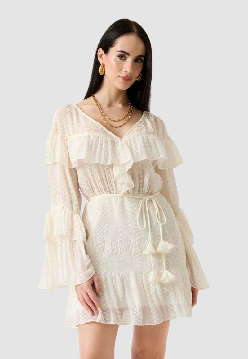 Femme aux longs cheveux foncés portant une robe crème à volants avec manches transparentes, une ceinture à nouer à la taille, des boucles d'oreilles en or et des colliers en or superposés.