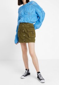 Maglione blu lavorato a maglia con motivo a foglia testurizzato, gonna cargo verde con bottoni frontali e tasche, e sneaker alte nere.