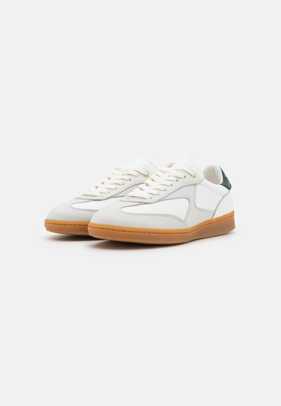 White leather sneakers with a textured finish, contrasting green heel accent, and tan rubber sole. Features flat white laces and stitching details.