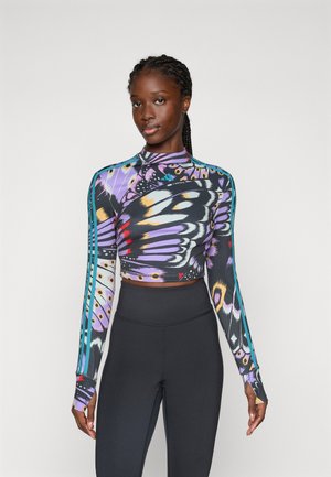 Ung kvinde med fletninger iført en langærmet crop top med farverigt sommerfuglevinge-mønster og sorte højtaljede leggings.
