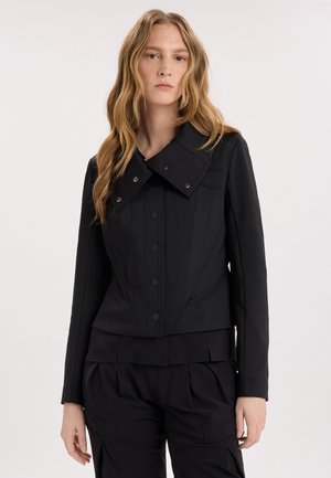 Donna con capelli lunghi e mossi che indossa una giacca nera aderente con dettagli a bottoni e pantaloni neri abbinati su uno sfondo chiaro e uniforme.
