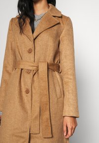 Donna che indossa un cappotto di lana color cammello con cintura sopra un maglione grigio, mostrando il torso e la mano destra su uno sfondo neutro.