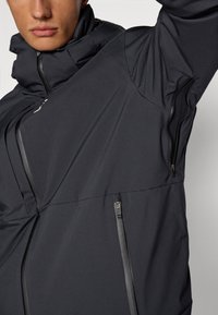 Schwarze Softshell-Jacke mit hohem Kragen, Reißverschlusstaschen und glatter Textur. Verfügt über ein tailliertes Design und eine stromlinienförmige Silhouette.