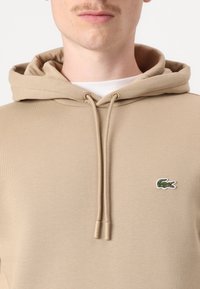 Homme portant un sweat à capuche beige avec des cordons de serrage et un petit logo de crocodile vert sur la poitrine, sur fond blanc.