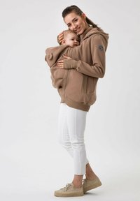 Sweat à capuche zippé marron avec une poche avant et une capuche. Il comporte une poche pour bébé à l'intérieur, associé à un pantalon blanc et des chaussures beiges.