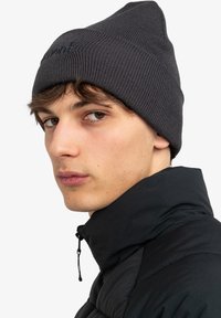 Beanie grigia a coste con punta e dettagli del logo in modo sottile, indossata con una giacca nera a collo alto, dotata di zip e tessuto testurizzato.