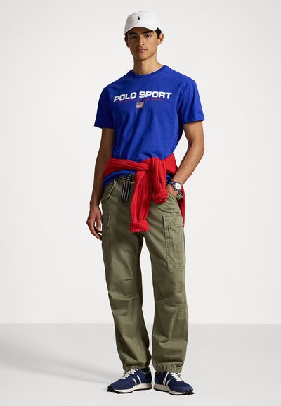 Modelo vistiendo una camiseta de algodón azul con el texto "POLO SPORT", pantalones cargo verde oliva, un suéter rojo atado alrededor de la cintura y zapatillas azules.