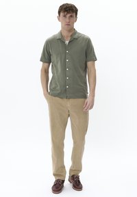 Camisa de botón de manga corta en color verde oliva, pantalones beige y zapatos marrones. La camisa tiene un collar y botones en la parte delantera.