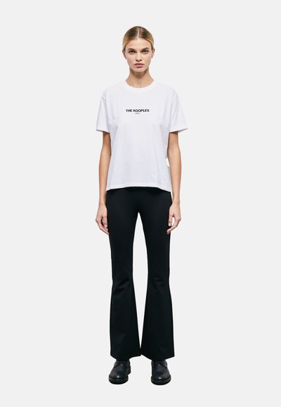 White cotton T-shirt with short sleeves, featuring black logo text "THE KOOPLES" centered on the chest. Paired with black flared trousers.