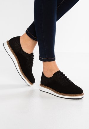 Czarne buty zamszowe z teksturowanym wzorem, białą środkową podeszwą i lekką platformą. Stylizowane z ciemnymi dżinsami, prezentujące casualowy wygląd.