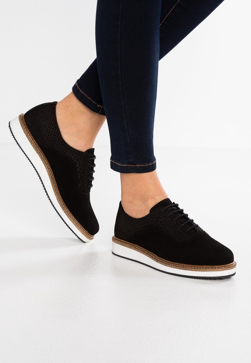 Chaussures en daim noir avec un design texturé, une semelle intermédiaire blanche et une légère plateforme. Portées avec un jean en denim foncé, affichant un look décontracté.